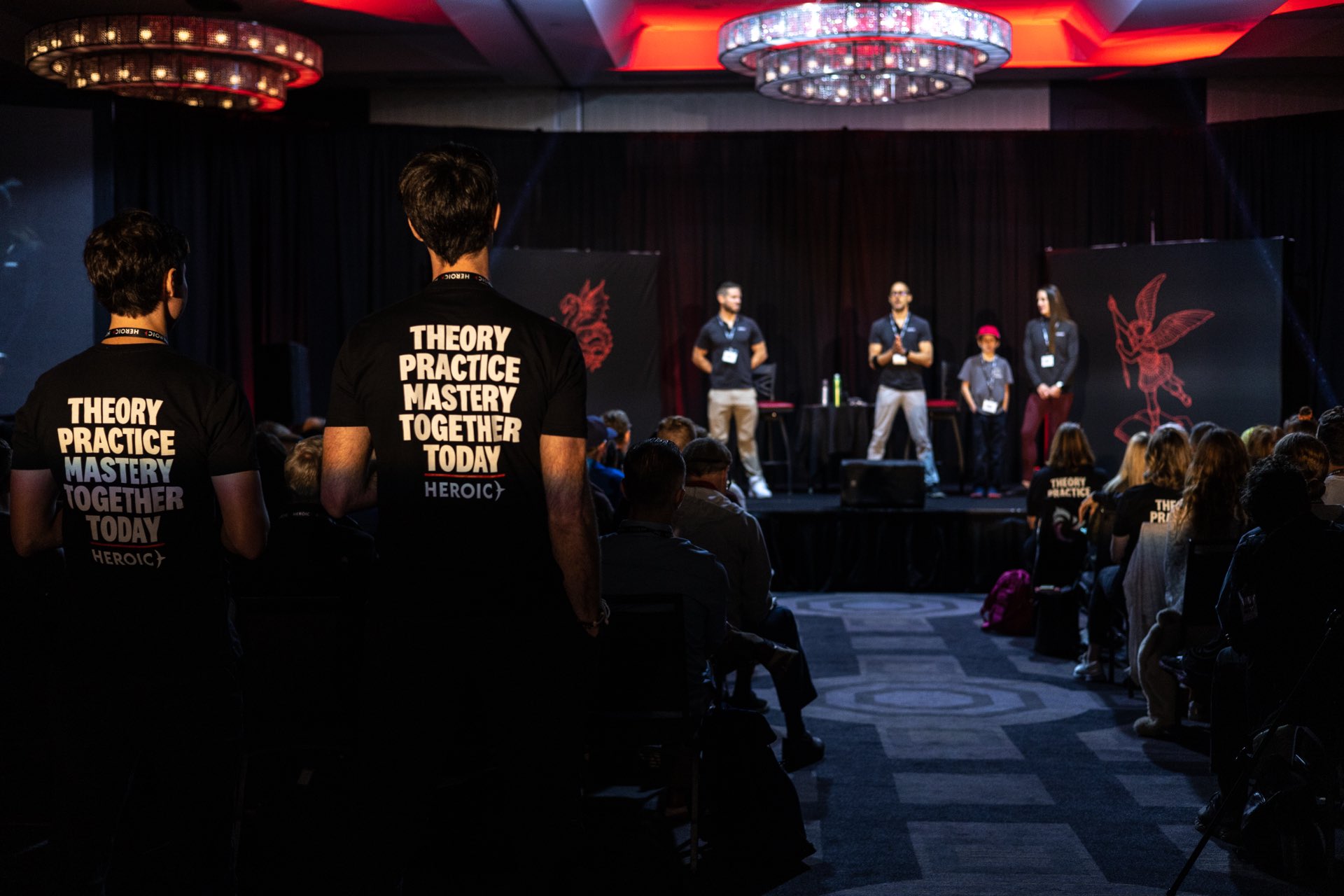 A Heroic event audience facing the stage, shirts reading Theory, Practice, Mastery, Together, Today